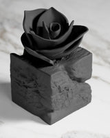Sculpture de rose noire jaillissant d'un socle en pierre brute, vue de face sur surface blanche