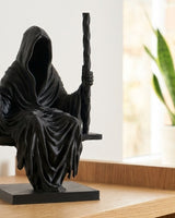 Sculpture de la Faucheuse en noir mat, assise sur une balançoire, vue de face