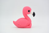 Figurine de flamant rose tricotée sur une étagère en bois à côté d'une petite plante et d'une déco neutre