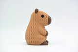 Petite figurine capybara façon tricot sur une étagère en bois avec plantes et livres, décor scandinave
